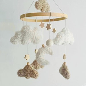 Op de afbeelding: Een baby mobile met een houten ring en hangende elementen. Het bevat pluizige witte en beige wolken, houten sterren, een maan en een bol. De elementen hangen aan touwtjes, wat een charmante decoratie creëert.
