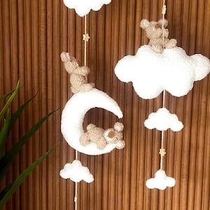 Könnte beinhalten: Ein skurriles Mobile für das Kinderzimmer mit flauschigen weißen Wolken, kleinen Sternen und entzückenden Teddybären. Ein Bär sitzt auf einem Mond, andere auf Wolken. Das Mobile hängt an Schnüren vor einem Holzgrund.