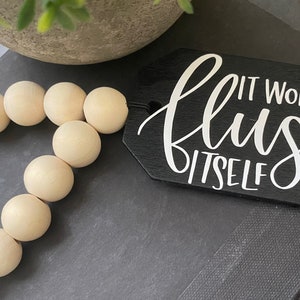 May include: A black wooden sign with white text that reads "It won't flush itself". The sign is hanging from a wooden bead garland.