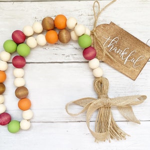 May include: A wooden bead garland with a mix of orange, red, green, and brown beads. The garland is tied with twine and has a burlap bow and tassel. A wooden tag with the word "thankful" is attached to the garland.