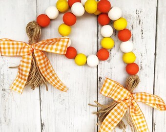 May include: A garland of wooden beads in orange, yellow, and white, with two gingham bows and jute tassels. The garland is perfect for fall decor.
