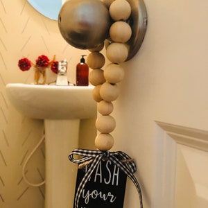 May include: A wooden bead garland with a black sign that reads "Wash your hands" hangs from a door knob. The sign has a black and white checkered ribbon tied around it.