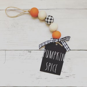 May include: A wooden bead garland with orange, white, and black and white checkered beads. The garland has a twine loop at the top and a black tag with the text "Pumpkin Spice" written in white.