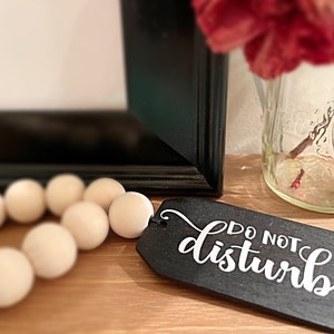 May include: A black wooden sign with white lettering that reads "Do Not Disturb". The sign is hanging from a strand of wooden beads.