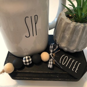 May include: A white ceramic mug with the word "SIP" printed on it, a small concrete planter with a succulent plant, and a black wooden sign with the word "COFFEE" printed on it. The items are arranged on a black wooden tray with a black and white checkered ribbon and wooden beads.