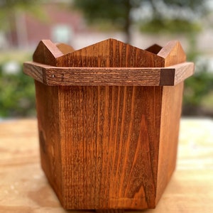 May include: A wooden planter with a hexagonal shape and a decorative top edge. The planter is made of dark brown wood and has a natural wood grain.
