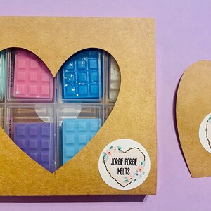 May include: A heart-shaped box made of brown cardboard with a cut-out heart shape revealing six wax melts in a variety of colours. The box has a sticker with the text "Jorge Porge Melts" and a floral design.