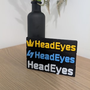 Puede incluir: Tres letreros rectangulares negros apilados sobre una superficie de madera. Cada letrero tiene el texto "HeadEyes" en un color diferente: amarillo, azul y blanco. Un jarrón negro con flores está en el fondo.