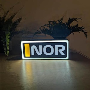 Puede incluir: Letrero rectangular iluminado con la palabra "NOR" en letras mayúsculas blancas. La letra "I" se reemplaza con un rectángulo vertical amarillo. El letrero está sobre un fondo oscuro con plantas frondosas.