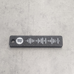 Könnte beinhalten: Ein schwarzer, rechteckiger Spotify-Code-Schlüsselanhänger mit strukturierter Oberfläche. Das Spotify-Logo befindet sich links, der Code ist weiß. Der Schlüsselanhänger hat oben ein kleines Loch zur Befestigung.