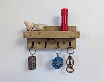 May include: A light brown wooden wall shelf with a small carved heart, featuring a top ledge holding two seashells and a red flashlight. Below the shelf are four black metal hooks, three of which hold keychains with various designs.