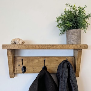 May include: A rustic wooden wall shelf featuring a flat top surface and a lower panel equipped with three black metal hooks. A small potted green plant sits on the shelf alongside a seashell. Two dark fabric items hang from the hooks.