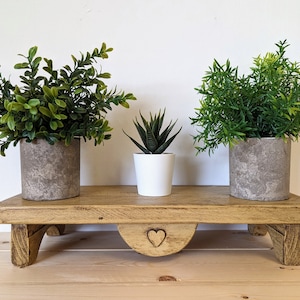 May include: Three potted plants on a wooden shelf with a heart-shaped cutout. The shelf has two legs on each side and is painted a light brown colour. The plants are in different coloured pots, one is white, one is grey, and one is a darker grey.