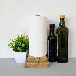 May include: A wooden paper towel holder with a square base and a small heart-shaped cutout on the front. The holder features a vertical rod supporting a roll of white paper towels. Next to it are a small green plant in a white pot and two bottles.