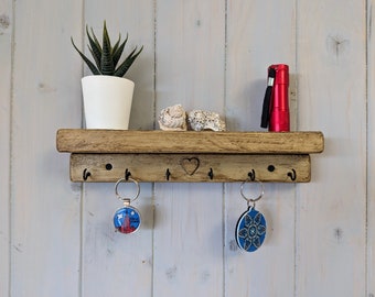May include: A wall-mounted wooden shelf with a natural finish featuring a carved heart in the center and five black metal hooks. Two keychains hang from the hooks, one with a New York graphic and one with a blue floral pattern.