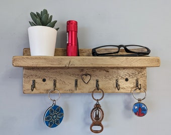 May include: A rectangular light brown wooden wall shelf with a carved heart in the center and six metal hooks. Three keychains hang from the hooks, featuring a blue mandala, a bronze bottle opener, and a round New York graphic.