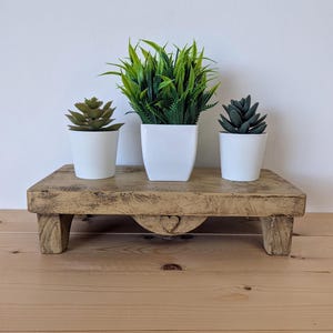 May include: A distressed wooden plant stand with three potted plants. The stand is rectangular with a heart-shaped cutout. The plants include a succulent, a tall green plant, and a dark green succulent, all in white pots. The stand is made of wood.