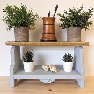 May include: A two-tiered wooden shelf, painted grey and brown. The top shelf displays two potted green plants and a wooden pen holder. The bottom shelf holds two small potted succulents and two small rocks.