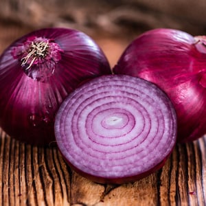 May include: Three red onions, one sliced in half, on a wooden surface. The sliced onion reveals the purple layers of the vegetable.