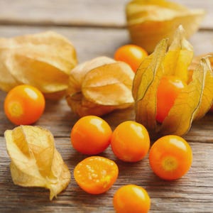 May include: Close-up of several orange physalis fruits, some still encased in their papery husks. The fruits are scattered on a weathered wooden surface, with one cut open to reveal its seeds. The husks are a light tan color.