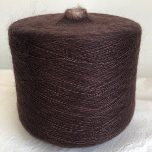 May include: A large spool of dark brown yarn. The yarn is tightly wound, creating a cylindrical shape. The top of the spool has a small, lighter-colored core. The yarn appears soft and fluffy, ideal for knitting or crocheting projects.