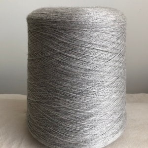May include: A large spool of gray yarn. The yarn is tightly wound around a cylindrical core, creating a dense, textured surface. The color is a neutral gray, suitable for various knitting or crochet projects. The yarn appears soft and fine.