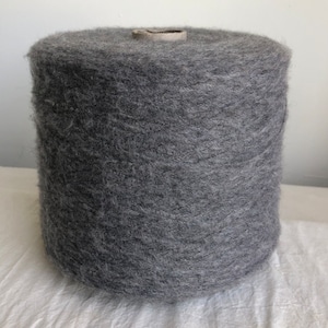 May include: A large, cylindrical spool of gray yarn. The yarn is tightly wound, with a slightly textured surface. A cardboard core is visible at the top. The yarn appears soft and fluffy, suitable for knitting or crocheting projects.