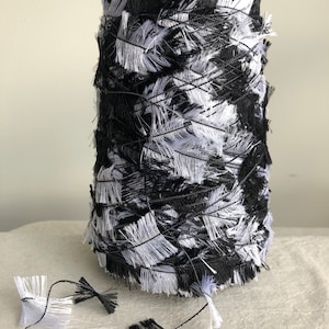 May include: A skein of black and white yarn with a fuzzy texture. The yarn is wound into a ball shape and is sitting on a white surface.
