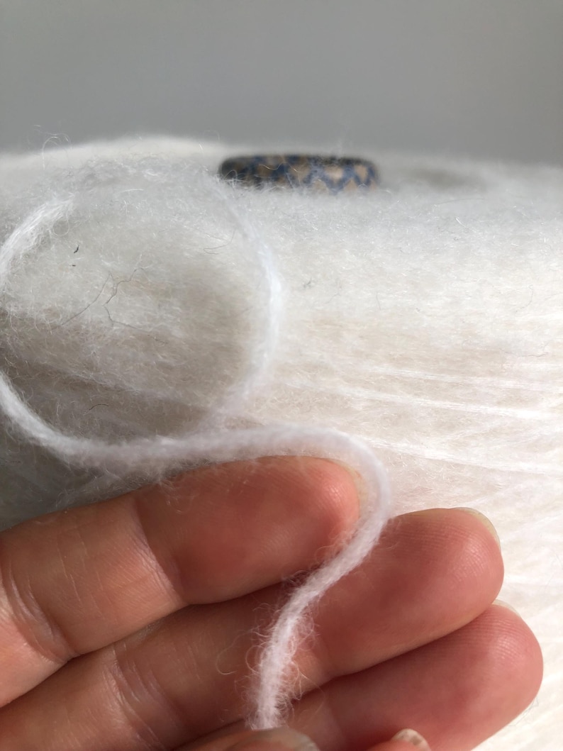 May include: A close-up of a white yarn skein with a single strand of yarn pulled out. The yarn is soft and fluffy, and the skein is wrapped in a white paper label.