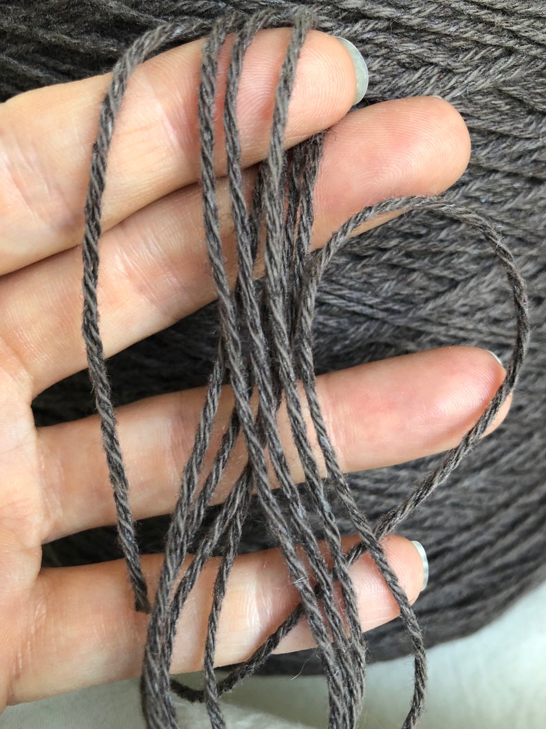 May include: Close-up of a dark grey yarn held in a hand. The yarn is twisted and has a subtle sheen.