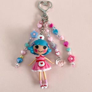 May include: A charming keychain with a doll in a pink dress and blue hair. The keychain has a heart-shaped clasp, beaded strands with hearts, flowers, and a clear charm. The doll has rosy cheeks and a heart on its dress.
