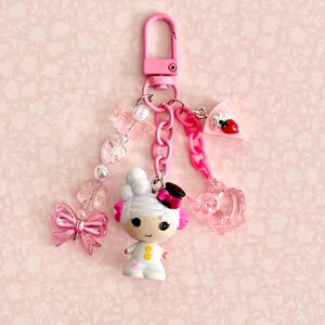 May include: A pink keychain with a lobster clasp, adorned with charms. Charms include a doll figure, a bow, a slice of cake with a strawberry, a clear elephant, and a beaded strand with butterflies and hearts.