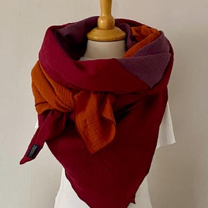 Könnte beinhalten: Ein dreieckiger Schal in den Farben Burgunderrot, Orange und Lila. Der Schal ist aus einem weichen, strukturierten Stoff gefertigt und um den Hals einer Schaufensterpuppe gefaltet und gebunden.