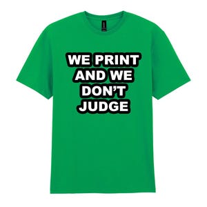 May include: A vibrant green t-shirt featuring the slogan "WE PRINT AND WE DON'T JUDGE" in bold white text with a black border. The tee has a crew neck and short sleeves.