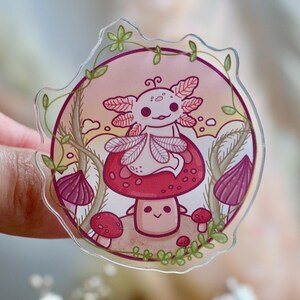 Könnte beinhalten: Ein klarer Aufkleber mit einem Cartoon-Axolotl, der auf einem roten Pilz mit weißen Punkten sitzt. Der Axolotl hat rosa Kiemen und ein lächelndes Gesicht. Der Aufkleber ist von einem rosa-grünen Blumendesign umgeben.