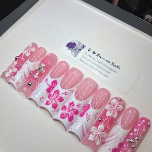 May include: A set of pink and white press-on nails with floral designs and rhinestones. Some nails are solid pink, while others feature hibiscus flowers and dot patterns. The nails are displayed in a white box with a business card.