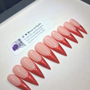 May include: Set of ten stiletto-shaped press-on nails. Each nail features a light pink base with a shimmering red French tip. The nails are arranged in a row on a white surface. A sticker with text and a skull graphic is visible.