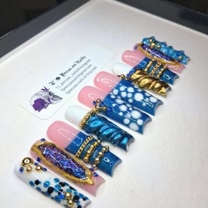 May include: A set of press-on nails in a white box. The nails feature various designs, including blue and white polka dots, gold accents, and jewel embellishments. Some nails have a pink base with white tips. The text "Tpresson Nails" is visible.
