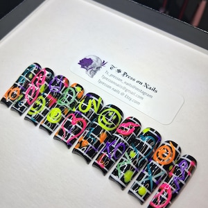 May include: Set of press-on nails with a black base, white grid pattern, and neon-colored smiley faces, hearts, and abstract designs. The nails are arranged in a row on a white surface. The text "Ts Press on Nails" is visible.