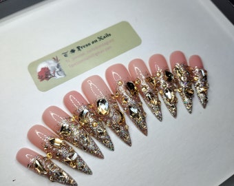 May include: A set of ten stiletto-shaped press-on nails. Each nail features a nude base with glitter and gold and silver embellishments, including rhinestones and metallic accents. The nails are displayed in a black box.