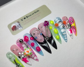 May include: A set of ten long, almond-shaped press-on nails with various designs. The nails feature a mix of colors, including pink, black, blue, and green, with embellishments like dice, studs, and floral accents. The nails are displayed in a white box.