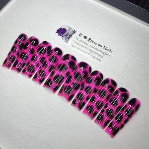 May include: A set of press-on nails with a vibrant pink base and black leopard print design. The nails are arranged on a white surface, with a sticker that reads "Press on Nails" and contact information.