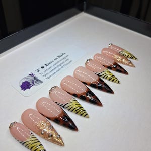 May include: A set of stiletto-shaped press-on nails. The nails feature a nude base with a French tip design. Some nails have a gold glitter accent, while others showcase a tiger stripe pattern and a tortoiseshell design. The brand name "Tpressonnails" is visible.