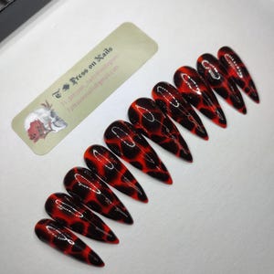 May include: Set of long, pointed press-on nails with a glossy finish. The nails feature a red and black pattern, resembling a fiery design. A small label with text is visible in the image.