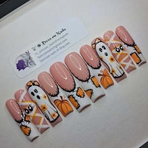 May include: A set of press-on nails with a light pink base and Halloween-themed designs. Each nail features a different design, including ghosts, pumpkins, and stitched patterns in orange, white, and black. The nails are arranged in a row.