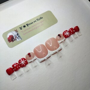 May include: Set of ten press-on nails with a white base and red and white snowflake designs. Some nails have a red accent with a small rhinestone. The nails are displayed on a white surface with a business card for "TPress on Nails" in the background.