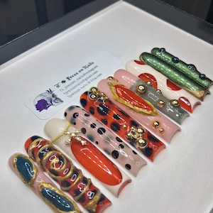 May include: A collection of press-on nails with various designs. The nails feature a range of colors, including red, black, gold, and green, with patterns like leopard print and abstract art. Some nails have gold embellishments and charms.