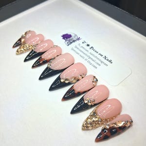 May include: Set of press-on nails with a light pink base, black tips, and gold glitter or rhinestone accents. Some nails feature a tortoiseshell design. The nails are displayed on a white surface.