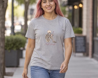 Puede incluir: Una camiseta gris jaspeado de manga corta con un gráfico de un esqueleto sentado sosteniendo una taza de café con un pequeño corazón. Debajo del esqueleto, el texto dice Still tired en letra cursiva. La camiseta tiene cuello redondo.