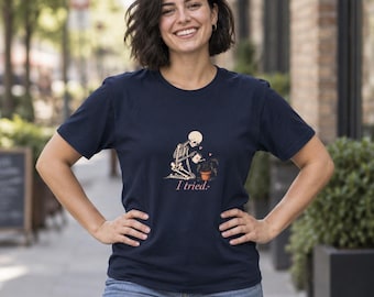 Puede incluir: Una camiseta de manga corta azul marino con un gráfico de un esqueleto arrodillado regando una planta en maceta con una pequeña regadera. Sobre la planta hay dos corazones pequeños y debajo del gráfico aparece el texto I tried.